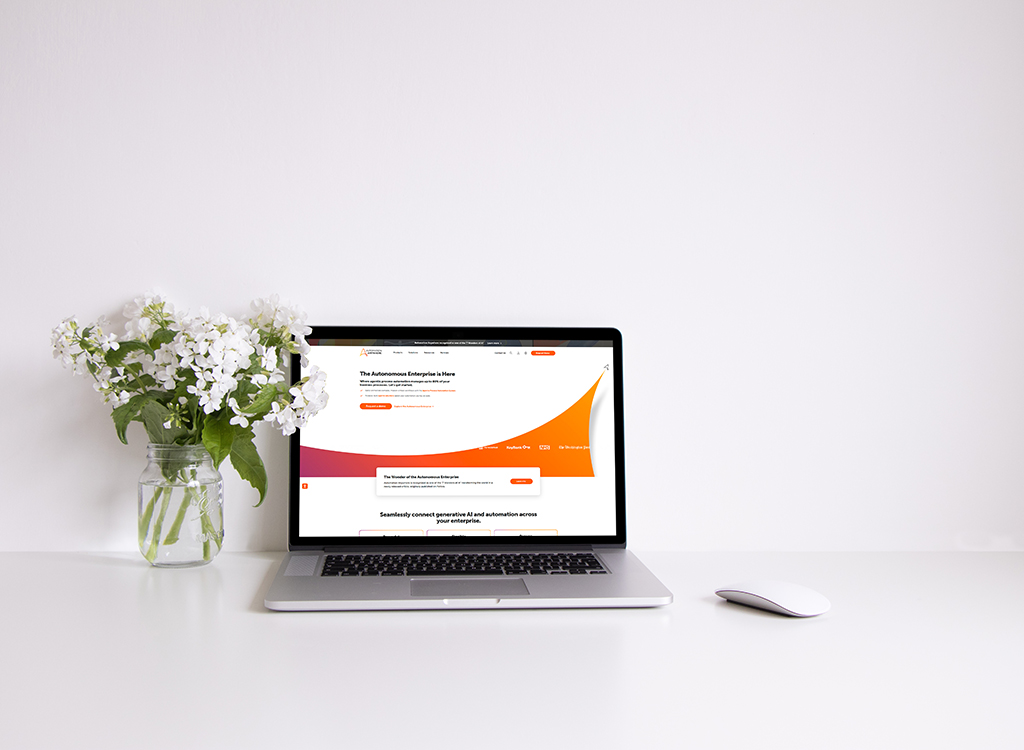 A modern workspace featuring a laptop displaying a website about autonomous enterprise solutions, accompanied by a vase of white flowers and a sleek mouse on a clean white desk.