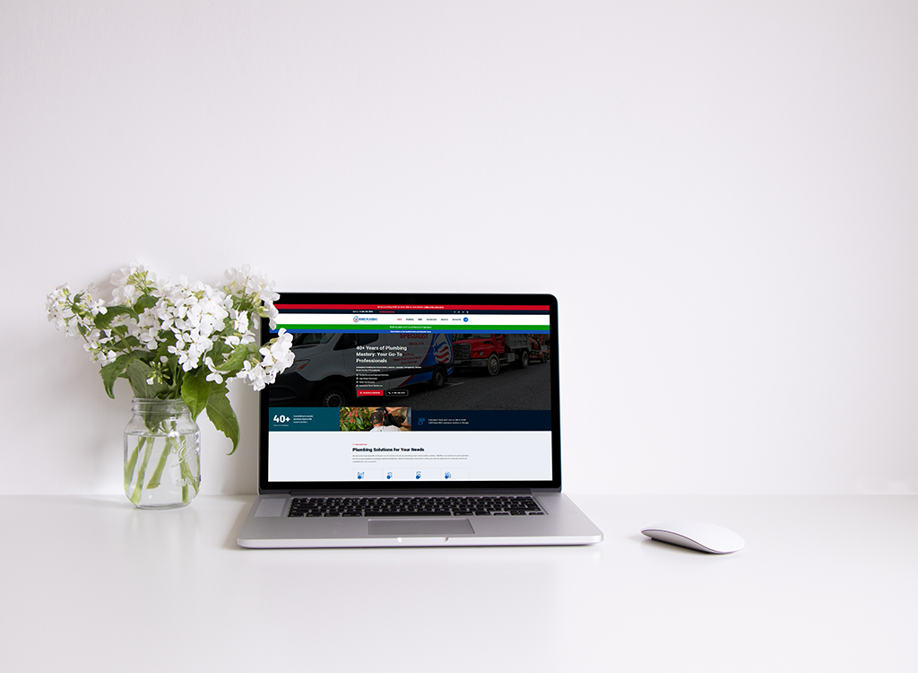 A silver laptop displaying a plumbing service website sits on a clean, white desk next to a vase of white flowers, illustrating a modern workspace setup.
