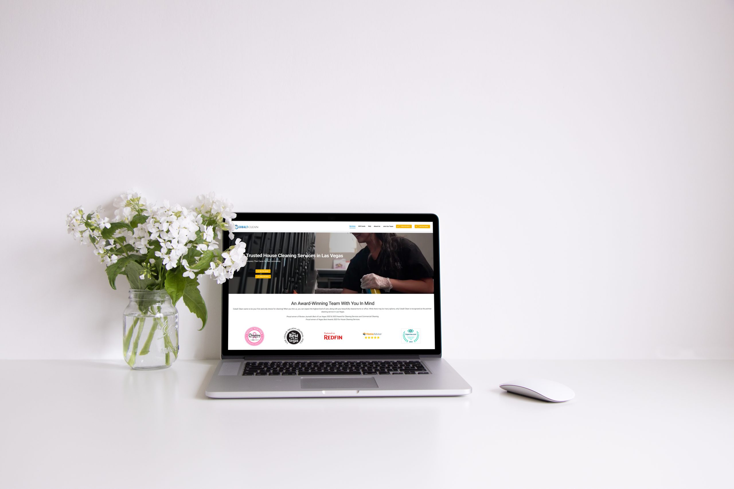 Laptop displaying a house cleaning service website on a white desk, accompanied by a vase of white flowers, emphasizing a clean and organized workspace.