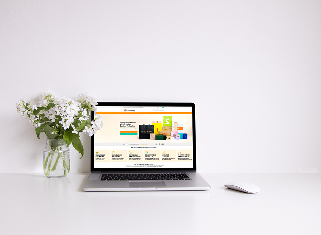 A modern laptop displaying a custom packaging website, surrounded by a vase of white flowers on a clean desk, emphasizing a professional and inviting workspace.