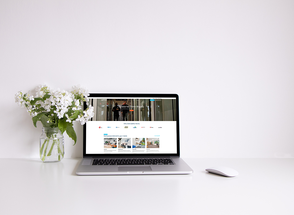 A modern laptop displaying a workspace website sits on a clean desk next to a glass vase filled with white flowers, creating a fresh and inviting home office atmosphere.