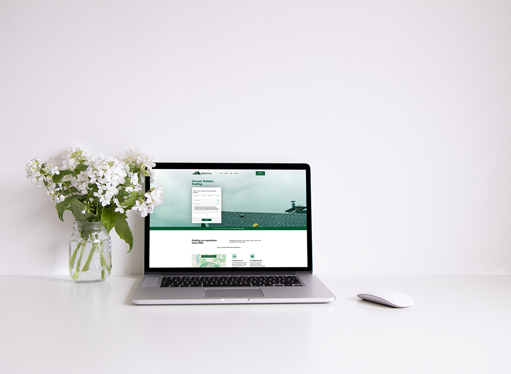 Laptop displaying a roofing company website with a green and white theme, surrounded by a vase of white flowers on a clean desk. The screen features a contact form and information about reliable roofing services.