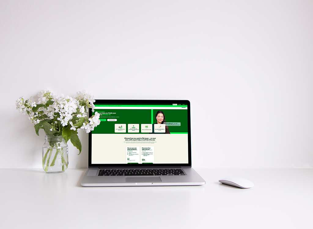 A laptop displaying a user-friendly website with a smiling woman on the homepage, surrounded by a vase of white flowers on a minimalist desk. The website features options for completing tasks and user assistance, emphasizing accessibility and support.