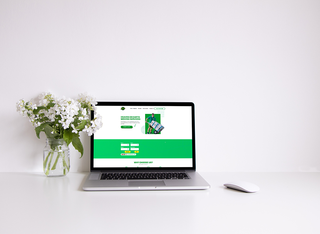 A laptop displaying a vibrant website for "Heaven on Earth Moving Services," featuring a green and white color scheme, sits on a clean white desk alongside a jar of fresh flowers and a mouse, emphasizing a professional and inviting workspace.