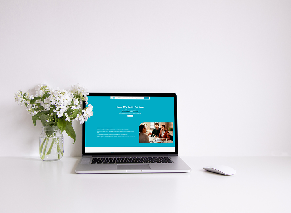 Laptop displaying a website about home affordability solutions, featuring a couple discussing financial options. A vase of white flowers is placed beside the laptop on a clean, minimalist desk.