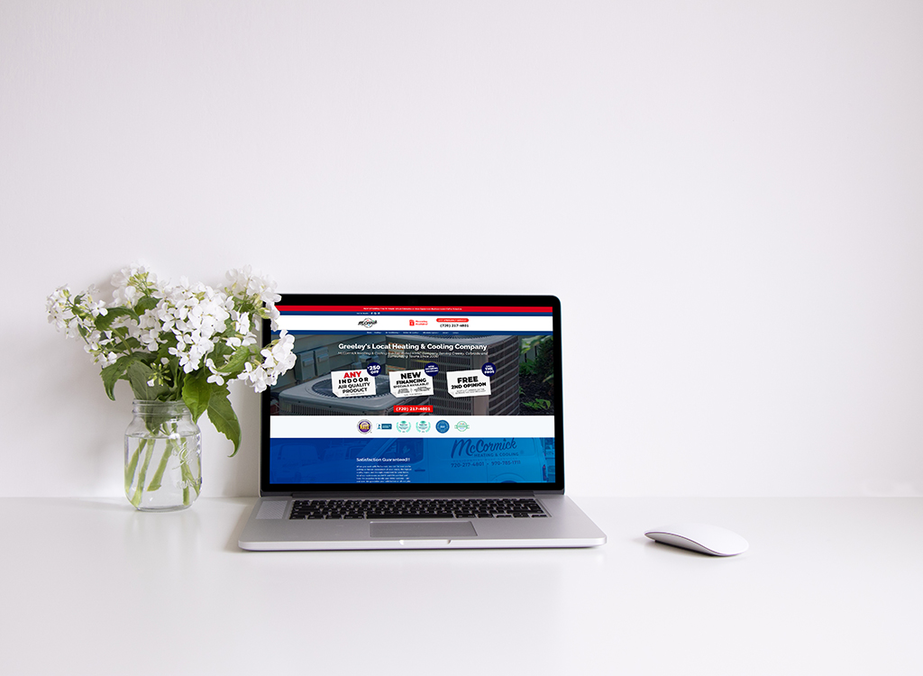 Laptop displaying the website of Greeley's local heating and cooling company, featuring promotional offers, contact information, and a clean workspace with a vase of flowers.