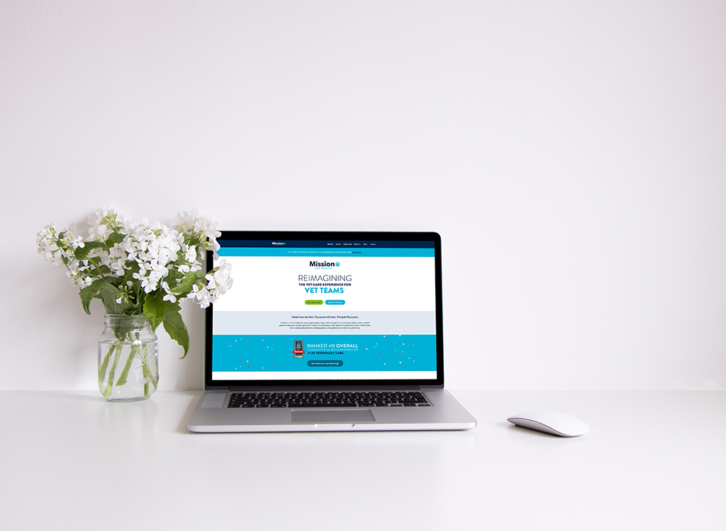A modern laptop displaying the "Mission" website, which focuses on reimagining the veterinary experience for vet teams, sits on a clean desk next to a glass vase of white flowers and a wireless mouse, emphasizing a professional workspace environment.