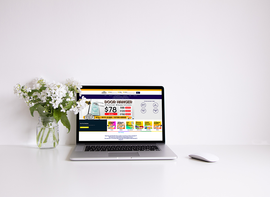 Laptop displaying a promotional webpage featuring a door hanger product priced at $78, alongside other printed materials, set on a clean white desk with a vase of white flowers.