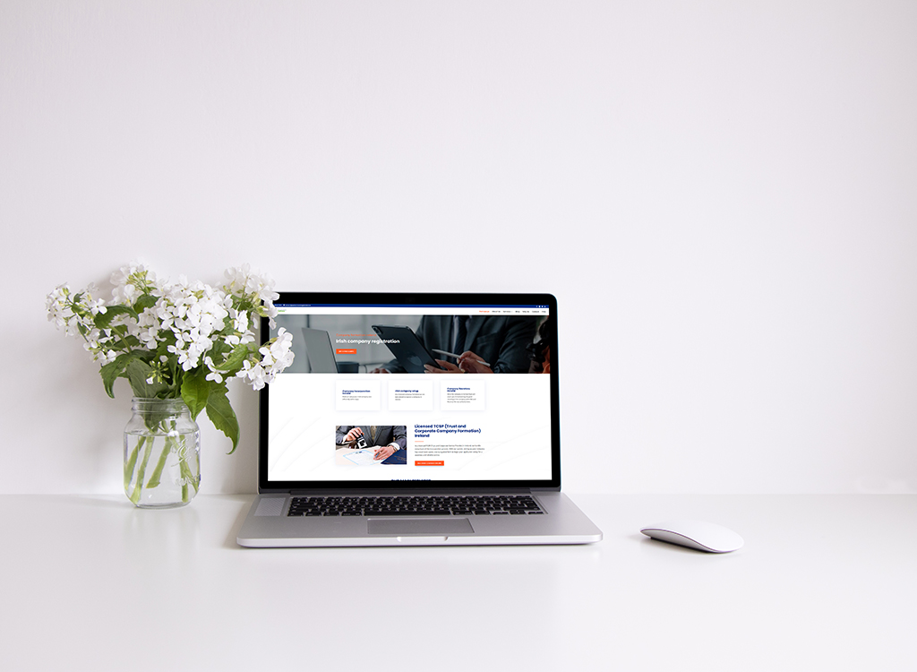 A modern workspace featuring a laptop displaying a website related to Irish company registration, accompanied by a white mouse and a vase of fresh flowers on a clean desk.