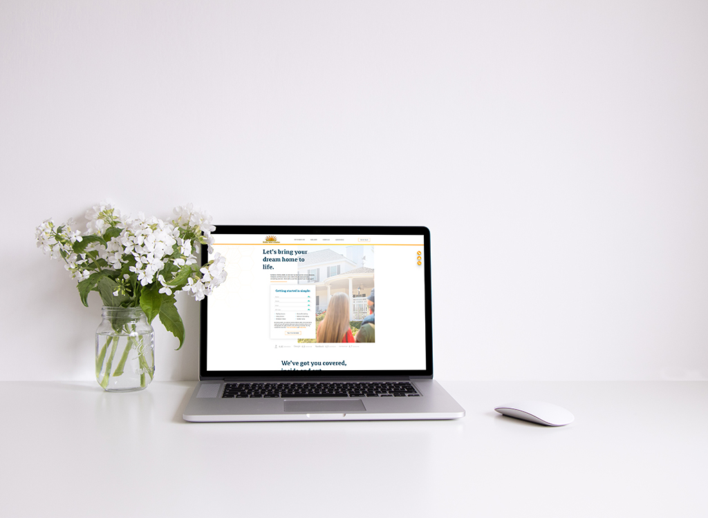 Laptop displaying a home design website on a white desk, accompanied by a vase of white flowers and a sleek computer mouse, emphasizing a clean and modern workspace aesthetic.