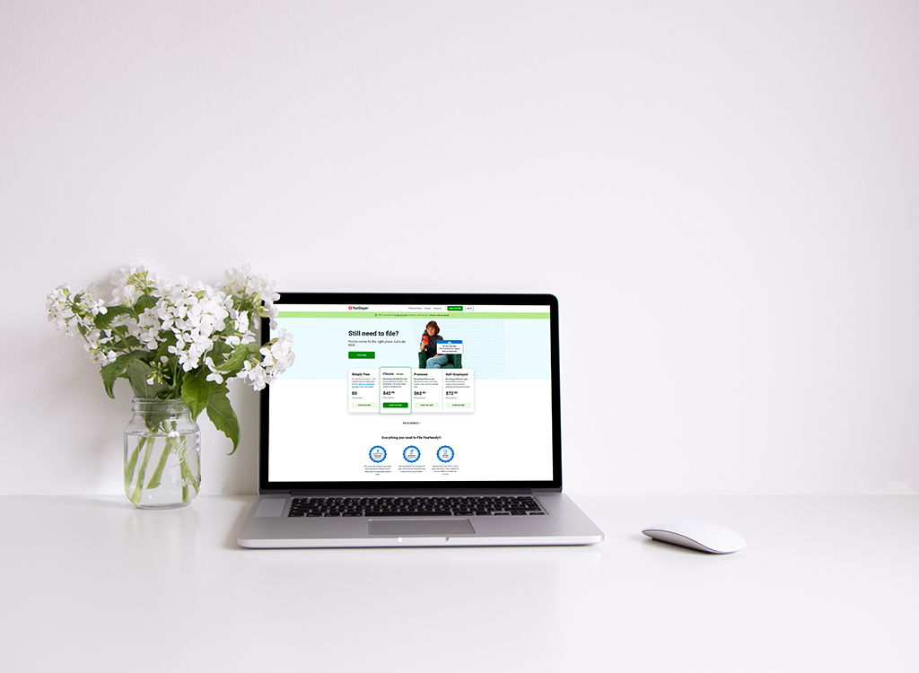 Laptop displaying a tax filing website with a clean interface, accompanied by a glass vase of white flowers on a minimalist desk. The scene suggests a professional and organized workspace for managing taxes online.
