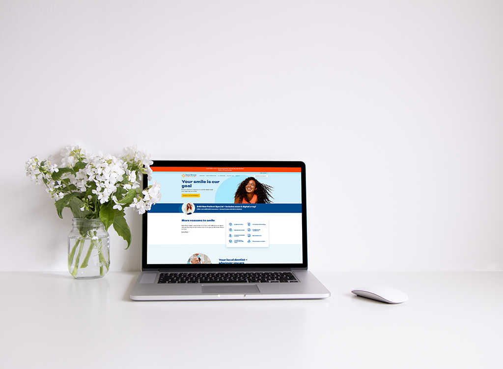 Laptop displaying a dental website focused on smile enhancement, placed on a clean desk with a vase of white flowers. The website highlights patient-centered dental services and features an inviting design.
