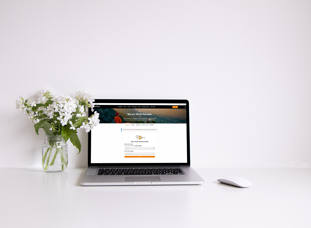 Laptop on a white desk displaying the World Nomads travel insurance website, accompanied by a glass vase of white flowers, illustrating a workspace setup for travelers.