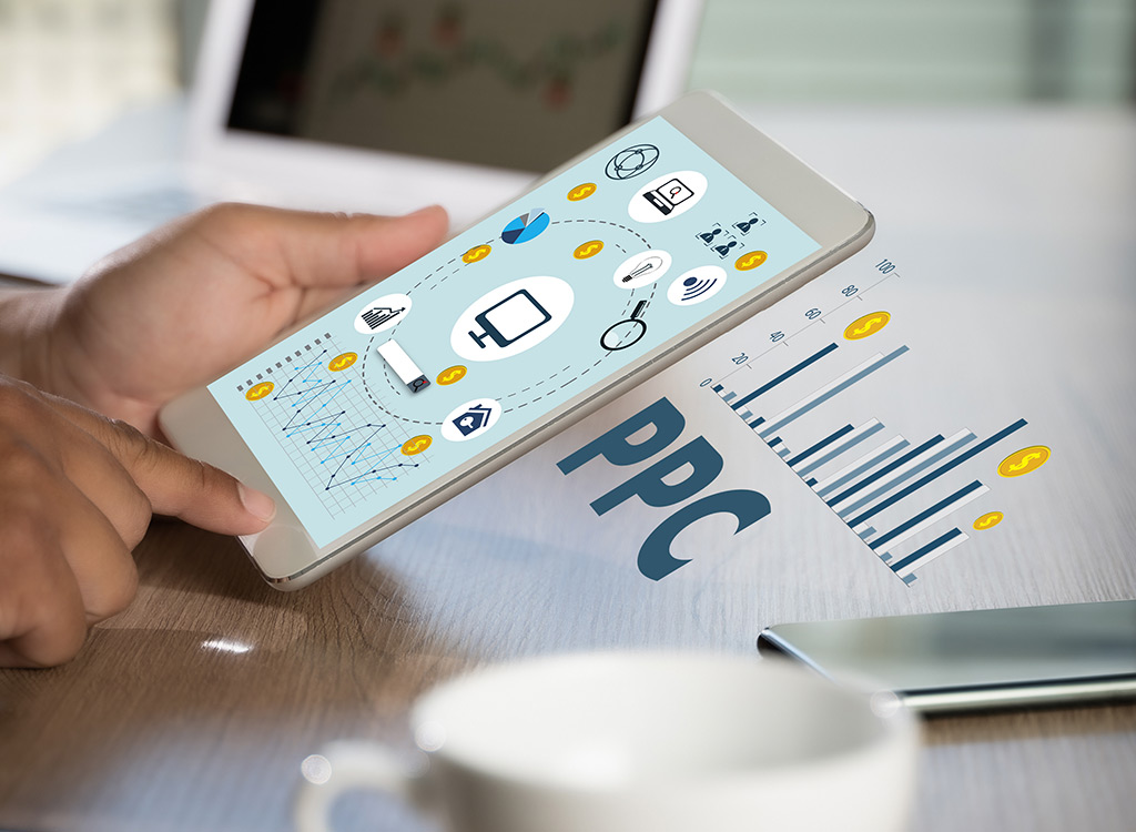 A person using a tablet displaying digital marketing analytics, featuring icons related to PPC (pay-per-click) advertising, graphs, and financial data, on a wooden desk with a coffee cup and smartphone in the background.